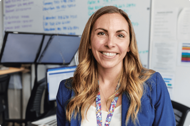 A young female staff member with long blond hair smiles as she stands in front of her desk.