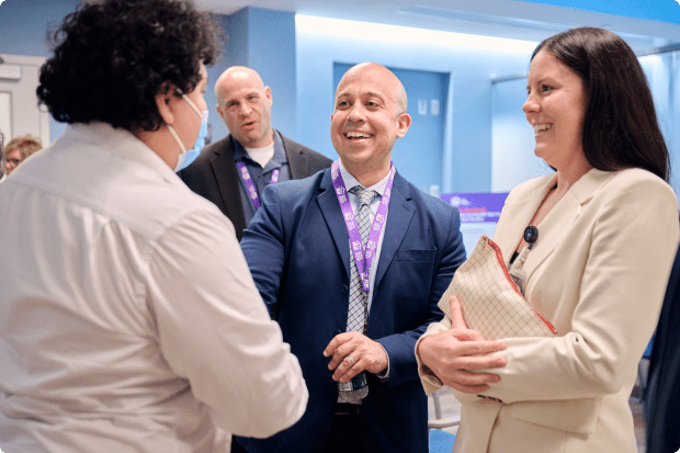 Three staff members in an office stand together smiling while one shakes hands with a visitor.