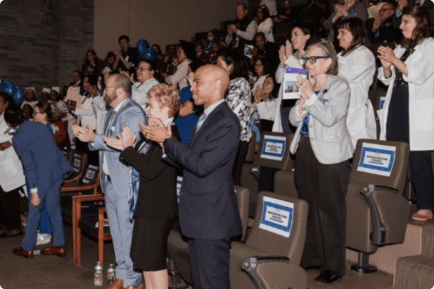 An audience of staff members stands and applauds at a staff appreciation event.