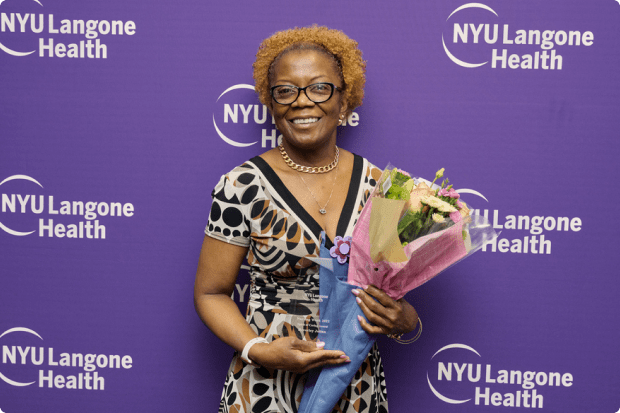 A female staff member smiles as she holds a bouquet while standing in front of a backdrop with the NYU Langone Health logo.