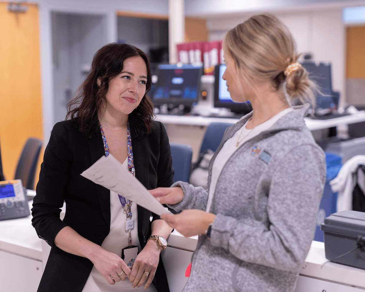 Two female staff at NYU Langone stand in a reception area and discuss information that one of them is holding.